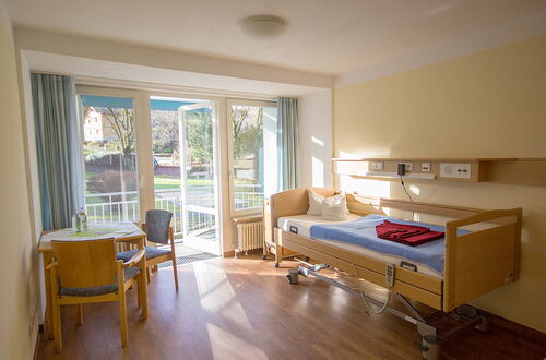 Ein helles Patientenzimmer mit einem verstellbaren Krankenhausbett, einem kleinen Tisch mit Stühlen und einer großen Fensterfront mit Blick ins Freie