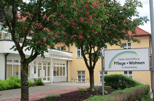 Eingangsbereich eines Gebäudes mit zwei blühenden Bäumen und einem Schild mit der Aufschrift 'Heilig-Geist-Stiftung Pflege + Wohnen in Nesselwang'